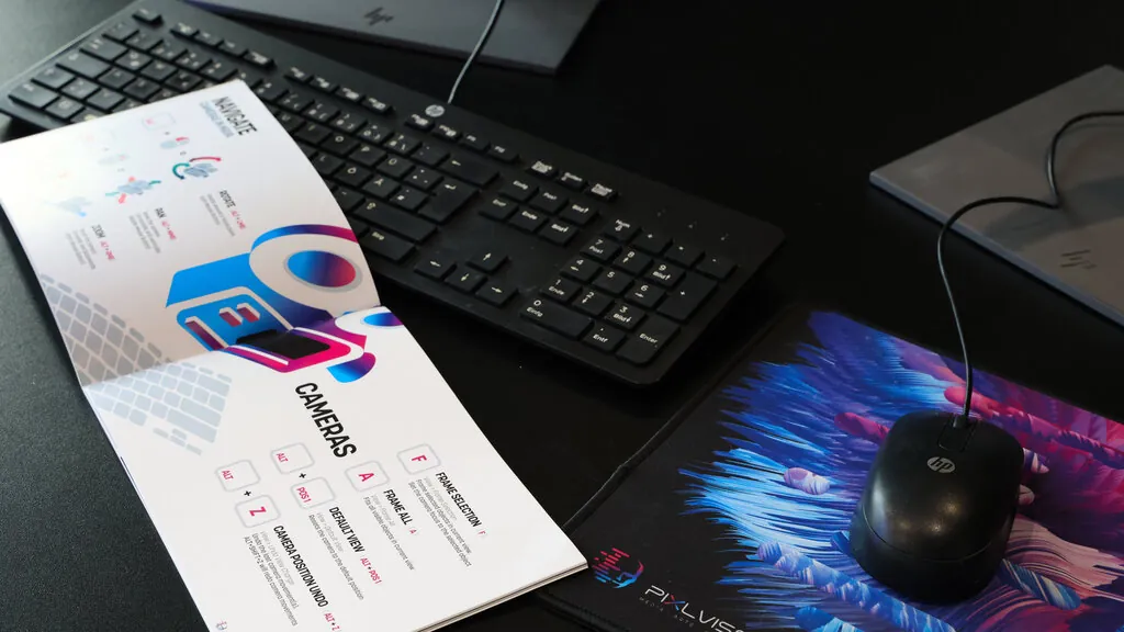 Close-up of a workspace featuring an HP keyboard, PIXL VISN mousepad, mouse, and an open booklet displaying colorful camera navigation tips.