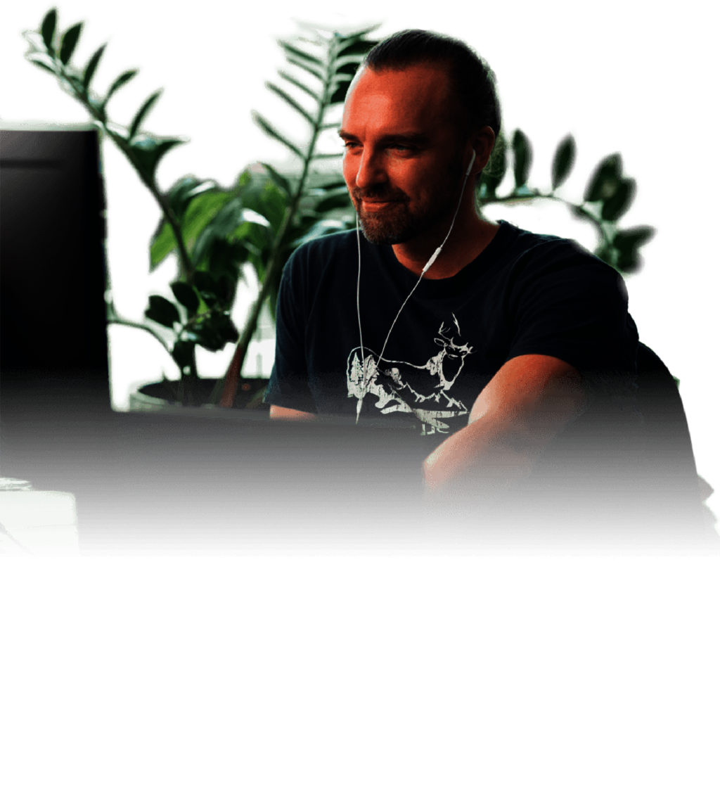 Portrait of a man with headphones at a screen – wearing a black T-shirt with an animal print, sitting in a room with a plant in the background. He has a focused, relaxed expression.